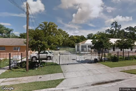 View of the entrance to a facility named Immaculee Home Care, showing a gated driveway with a black metal fence, a white vehicle parked inside, and two buildings surrounded by trees under a partly cloudy sky.