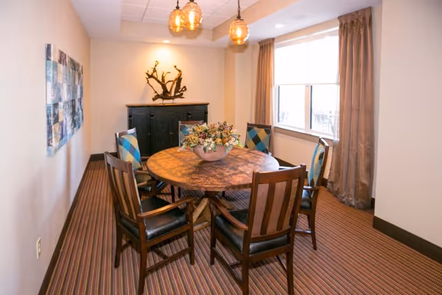 A small dining room with a round wooden table surrounded by six chairs, some with patterned cushions. A decorative flower arrangement is placed in the center of the table. The room has a striped carpet, beige walls, a window with brown curtains, and a black cabinet against the far wall with a decorative sculpture on top. Three pendant lights hang from the ceiling.