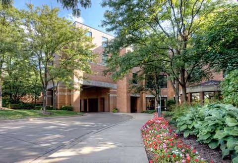 Brick senior living facility exterior with a curved driveway, trees, and flower beds.