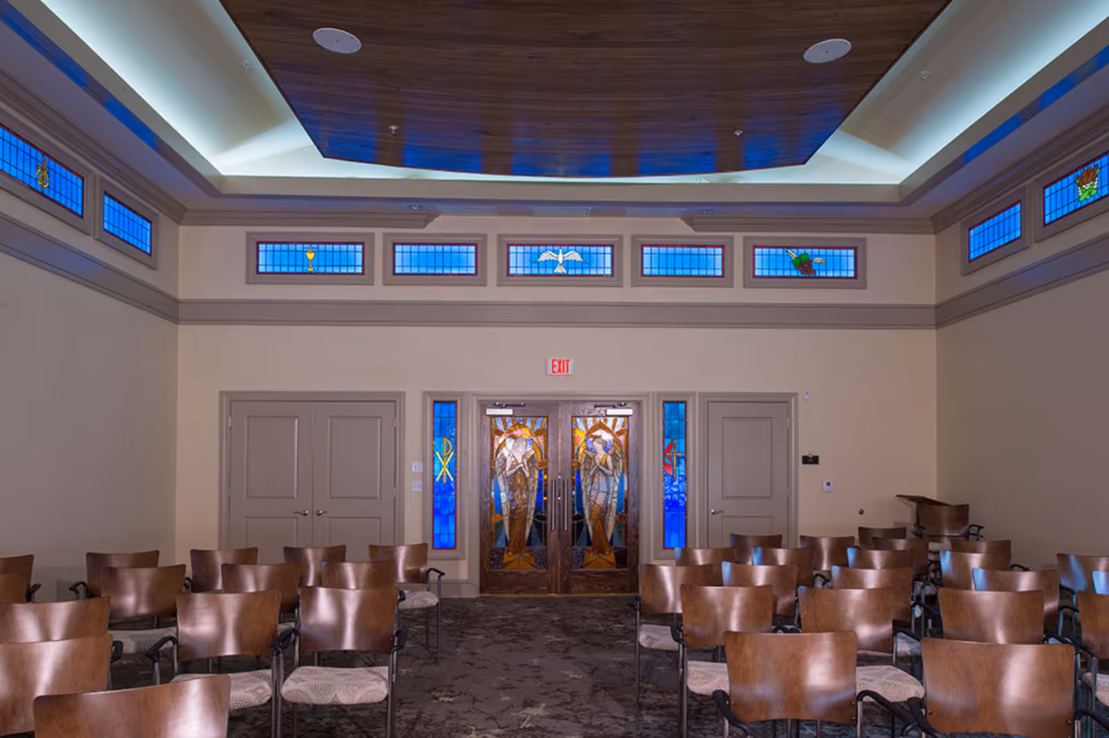 Interior meeting/chapel room with rows of wooden chairs facing stained-glass double doors and clerestory windows under a coffered ceiling.