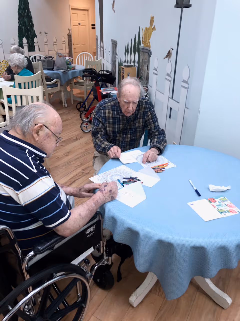 Two elderly men sitting at a round table covered with a blue tablecloth, engaged in coloring and drawing activities with crayons and paper. One man is in a wheelchair wearing a striped shirt, and the other is wearing a plaid shirt. In the background, there are more elderly people seated at tables, a walker, and a wall with decorative murals of animals and plants.