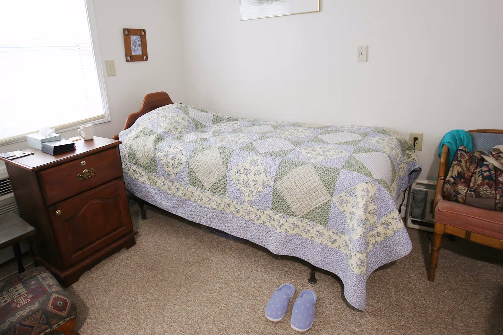 Simple single bedroom with a quilt-covered bed, wooden nightstand, chair, and slippers on the carpeted floor.