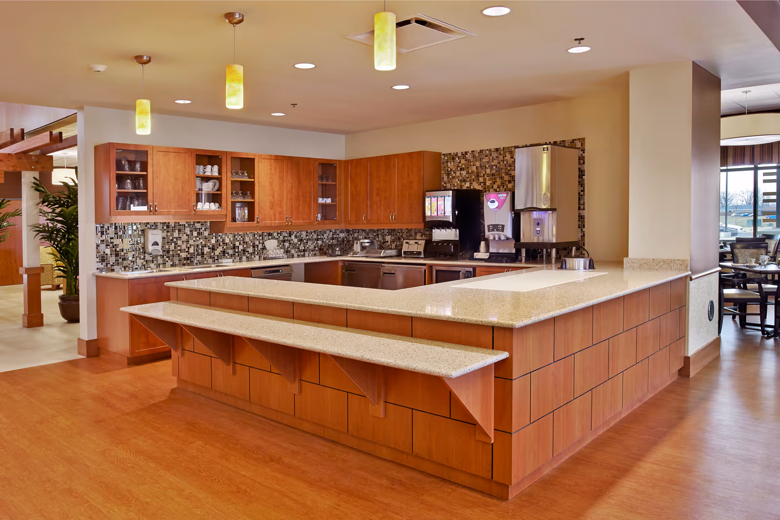 A modern kitchen area with wooden cabinets, a mosaic tile backsplash, and a large countertop island with a built-in bench. The kitchen includes a beverage dispenser and various kitchen appliances. Pendant lights hang from the ceiling, and there is a dining area visible in the background.