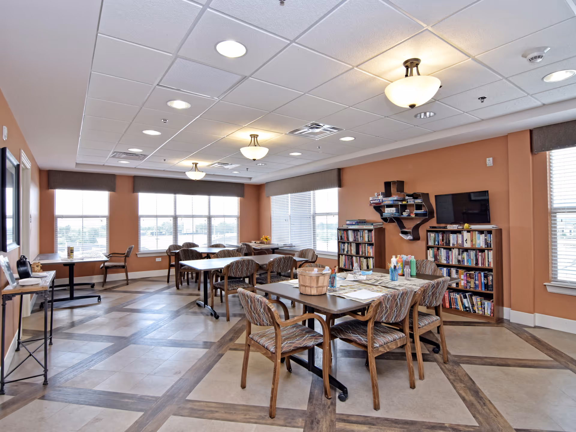 A well-lit common room with multiple tables and chairs arranged for group activities or dining. The room features large windows with blinds, a bookshelf filled with books, a wall-mounted TV, and a uniquely shaped wall shelf holding board games. The walls are painted a warm orange color, and the ceiling has recessed lighting and hanging light fixtures.