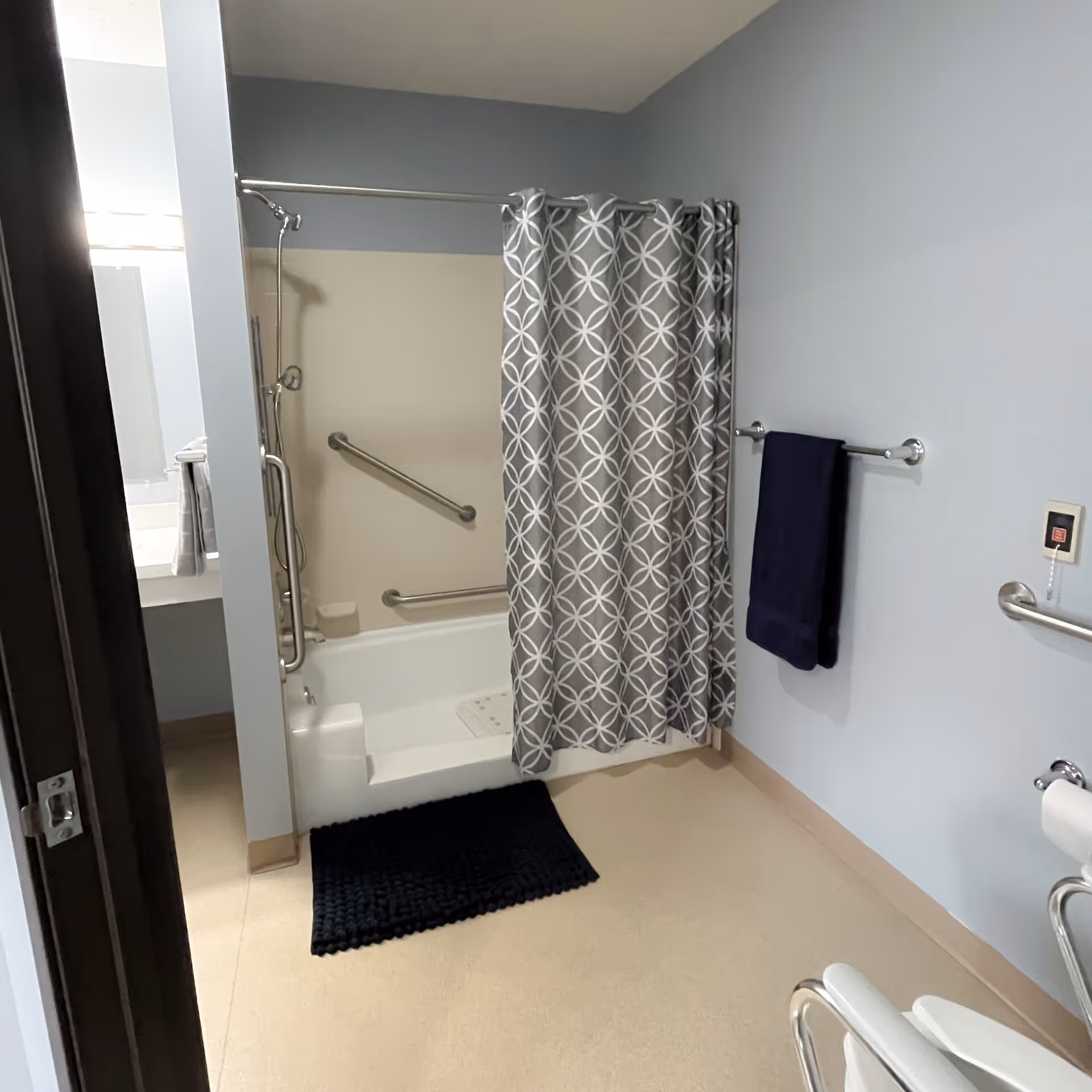 A bathroom with a bathtub and shower combination featuring a gray and white patterned shower curtain. There are grab bars installed on the wall inside the bathtub area and on the adjacent wall. A dark blue towel hangs on a towel rack, and a black bath mat is placed on the floor in front of the tub. The walls are painted light blue, and a toilet with support rails is partially visible on the right side.