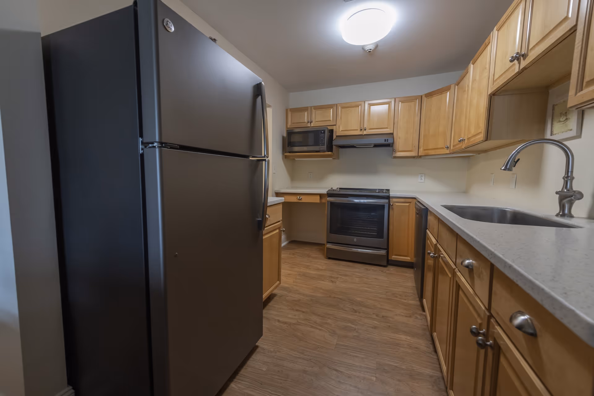 A kitchen with wooden cabinets, a stainless steel refrigerator, microwave, stove, dishwasher, and a sink with a modern faucet. The floor is wood, and the walls are painted white.