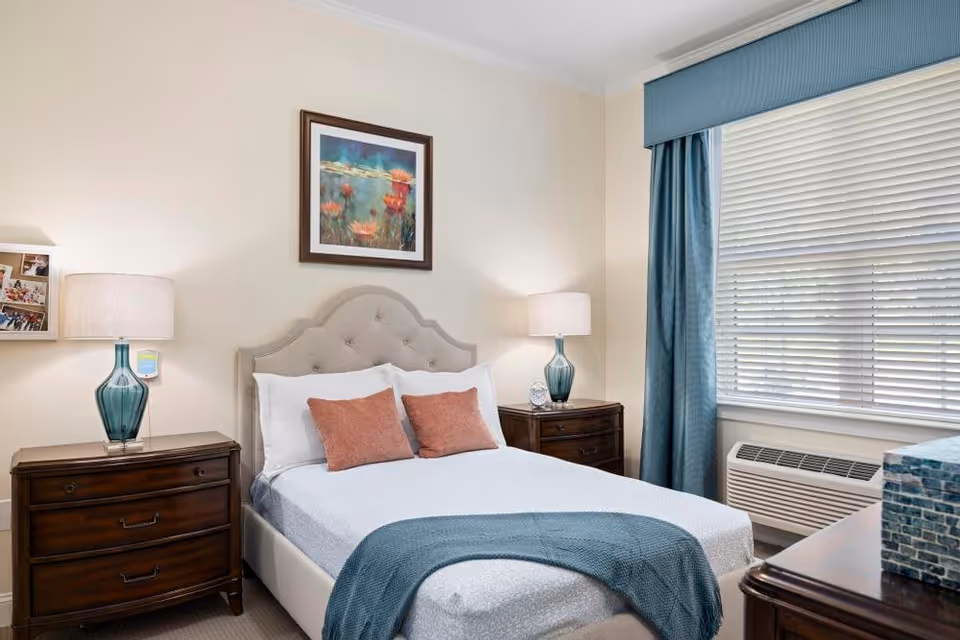 A neatly made bed with a tufted beige headboard, white bedding, two rust-colored pillows, and a blue throw blanket at the foot. On either side of the bed are dark wooden nightstands with blue glass lamps. A framed floral painting hangs above the bed. To the right, a window with white blinds and blue curtains lets in natural light. An air conditioning unit is below the window.