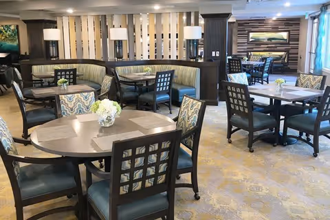 A spacious and well-lit dining area with multiple round and square tables surrounded by cushioned chairs. The room features a mix of patterned and solid upholstery, decorative lamps, and floral centerpieces on each table. The walls have a combination of wood paneling and light-colored accents, with a large framed landscape painting visible in the background.