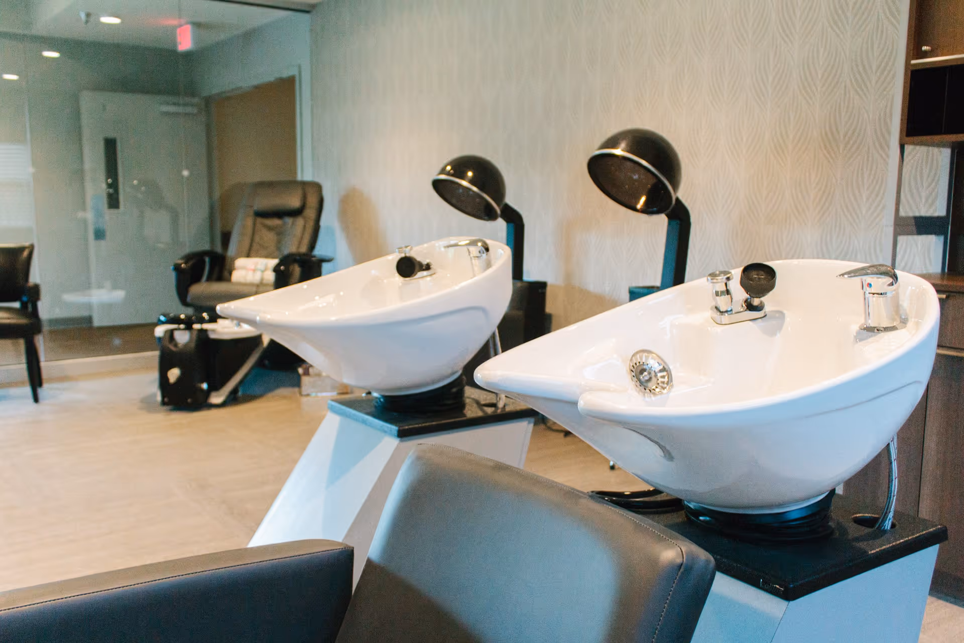 Interior view of a salon area in a senior living facility with two white hair washing sinks, black salon chairs, and two black hair dryers mounted on stands against a patterned wall.