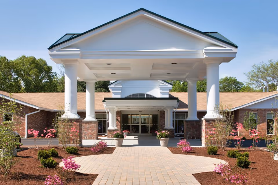 Front entrance of Kent Regency facility featuring a covered driveway with white columns, brick walls, and landscaped flower beds with pink flowers and green shrubs under a clear blue sky.