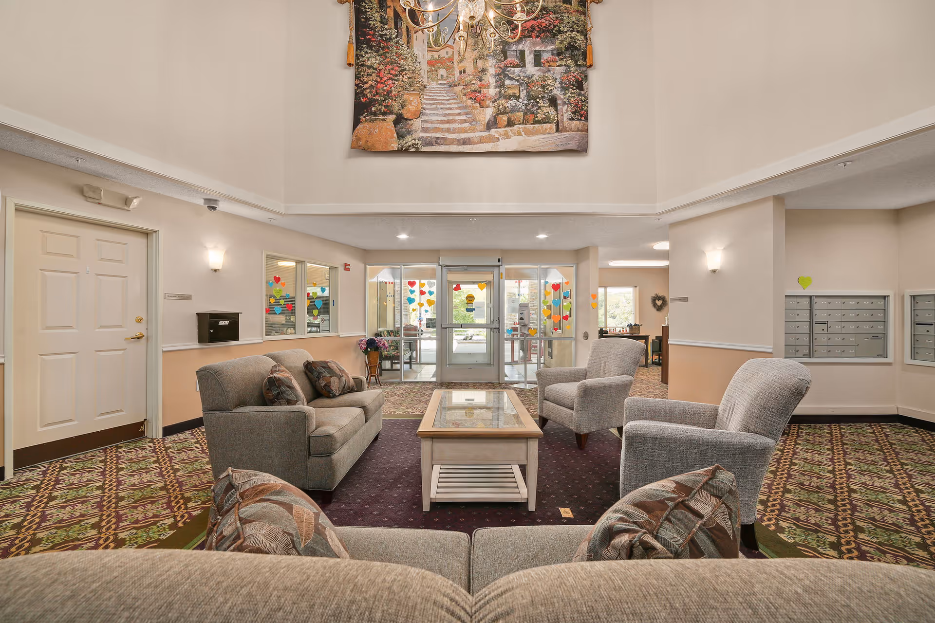 A spacious and well-lit lobby area with comfortable seating including sofas and armchairs arranged around a glass-top coffee table. The floor is carpeted with a patterned design, and there is a large decorative tapestry hanging on the wall above. The entrance features glass doors decorated with colorful heart-shaped stickers. Mailboxes are visible on the right wall, and there is soft wall lighting throughout the space.