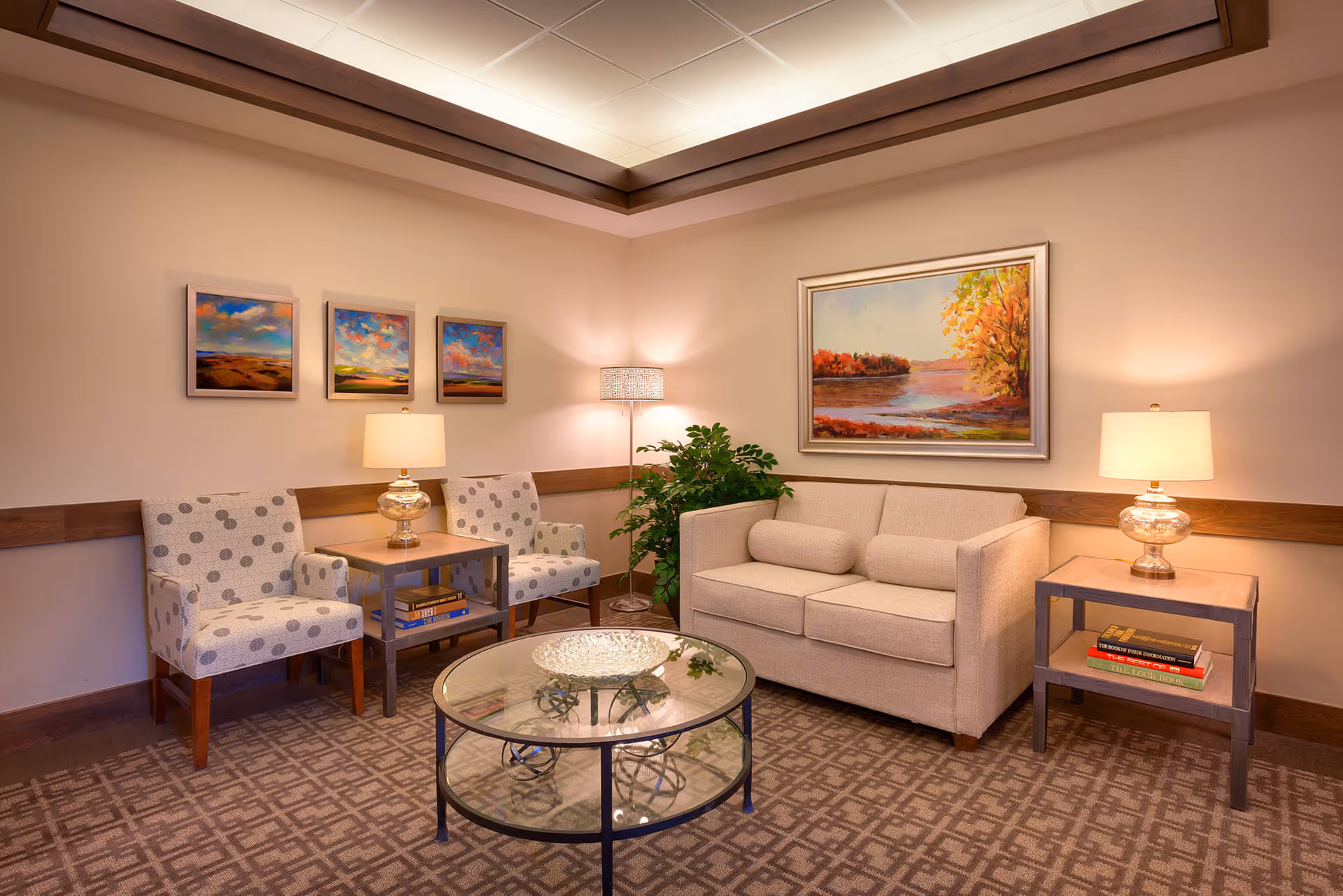 A cozy lounge with a sofa, two armchairs, table lamps, a glass coffee table, and framed artwork on the walls.