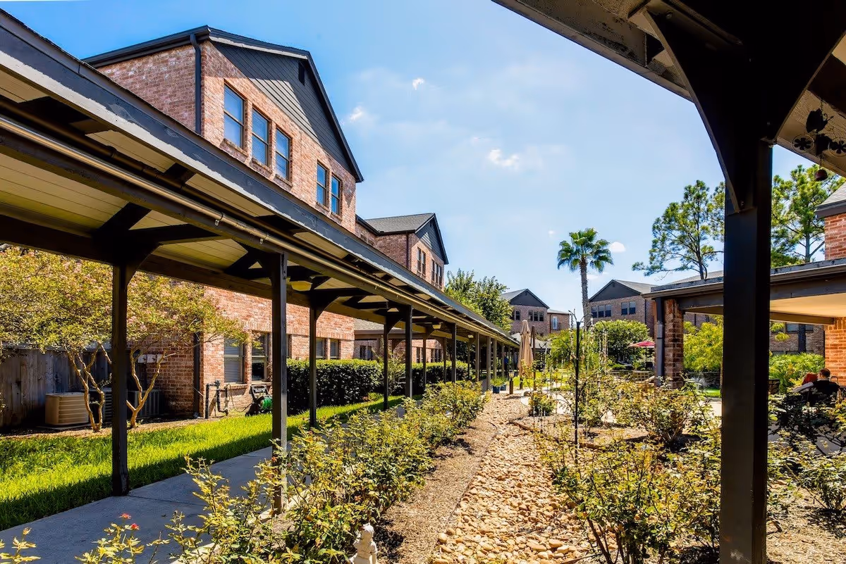 Outdoor view of Tarrytowne Estates showing a covered walkway with black support beams, landscaped garden with bushes and small trees, and brick buildings in the background under a clear blue sky.