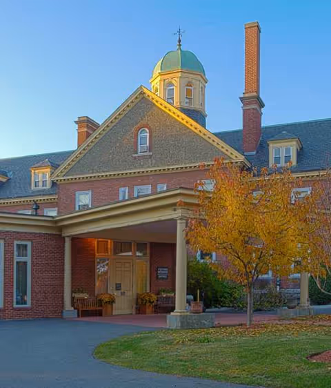 Front view of a brick senior living facility with a porte-cochère and autumn tree.