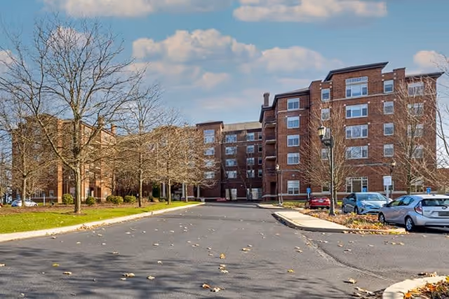Exterior view of a multi-story brick building with several windows, surrounded by leafless trees and parked cars under a partly cloudy sky.