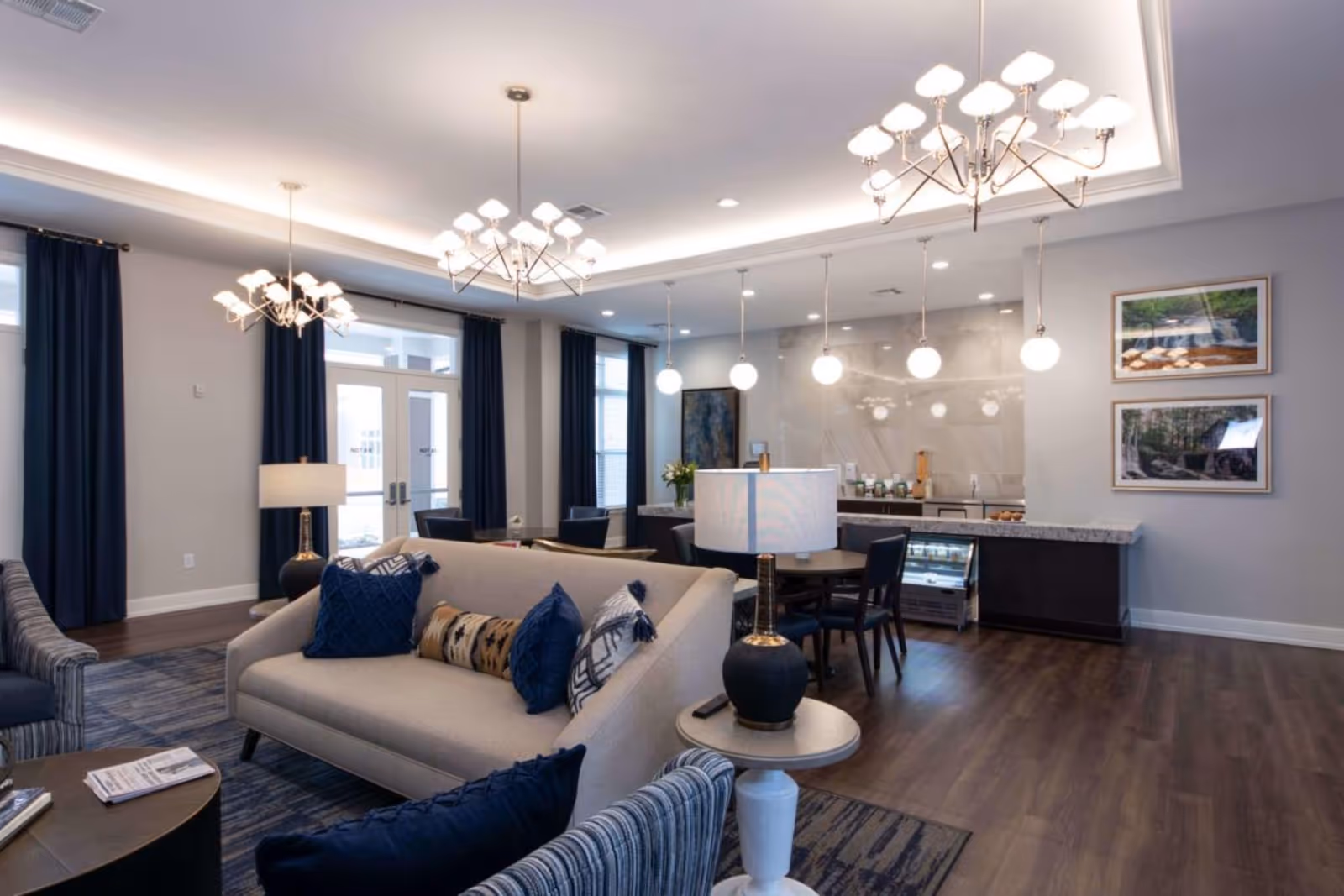 Bright, modern senior living common area with sofas, a coffee table, dining chairs, and a kitchenette under pendant chandeliers.