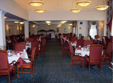 Spacious dining room with multiple round tables set with white tablecloths and red upholstered chairs under hanging ceiling lights.