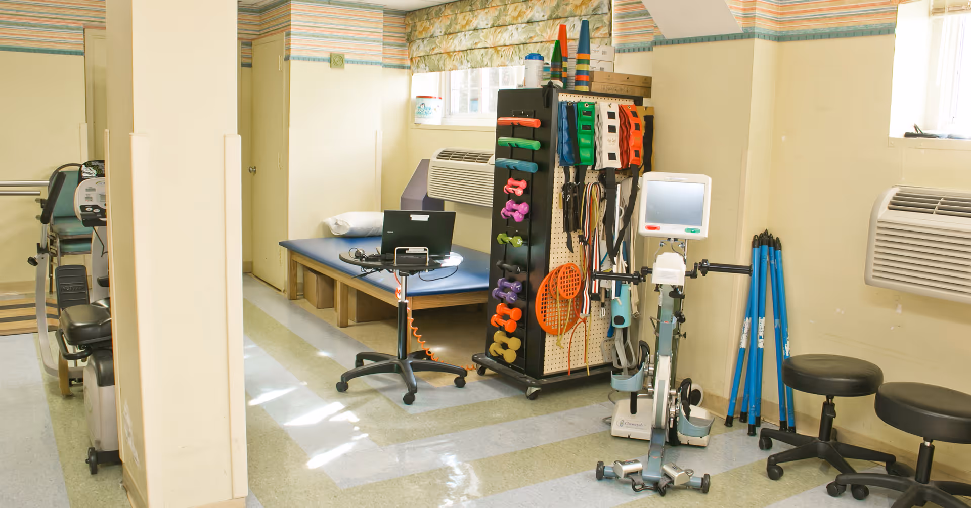 A physical therapy or rehabilitation room with exercise equipment including a stationary bike, colorful dumbbells, resistance bands, a treatment table with a pillow, and stools. The room has light yellow walls, windows with floral curtains, and air conditioning units.