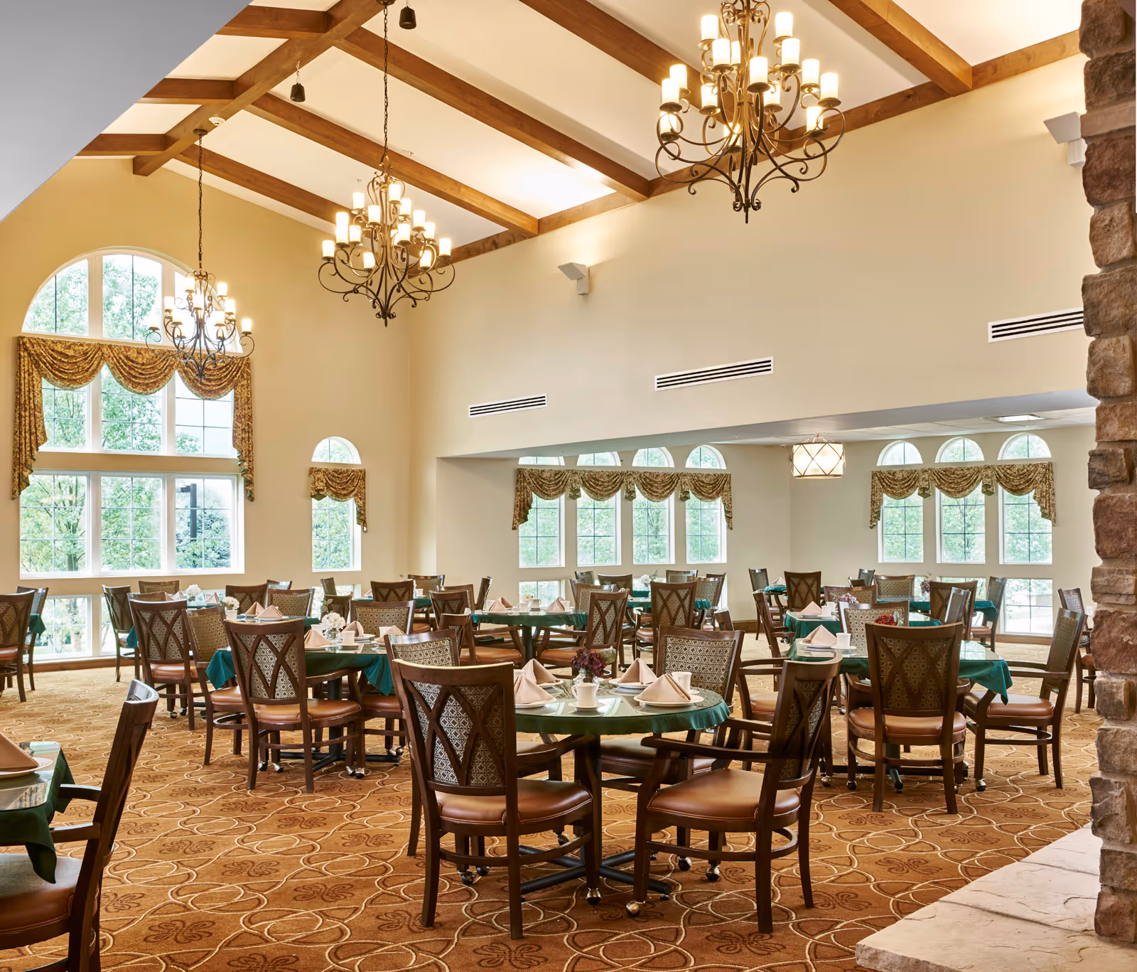 A spacious dining room with multiple round tables covered with green tablecloths and set with napkins and cups. The room features large windows with decorative curtains, high ceilings with wooden beams, and elegant chandeliers hanging from the ceiling. The carpet has a patterned design, and there is a stone fireplace partially visible on the right side.