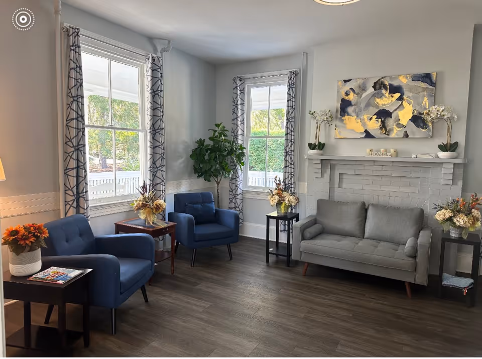 A cozy senior living community lounge with two blue armchairs and a gray sofa arranged around a white painted brick fireplace. The room has two large windows with patterned curtains, a wooden floor, and several floral arrangements on side tables. Above the fireplace hangs an abstract painting with gold, gray, and blue tones.