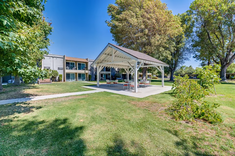 Outdoor area of Escondido Senior Living featuring a white pavilion with a pitched roof and several tables and chairs underneath. Surrounding the pavilion is a well-maintained grassy lawn with trees and shrubs, and in the background, there is a two-story building with balconies and large windows under a clear blue sky.