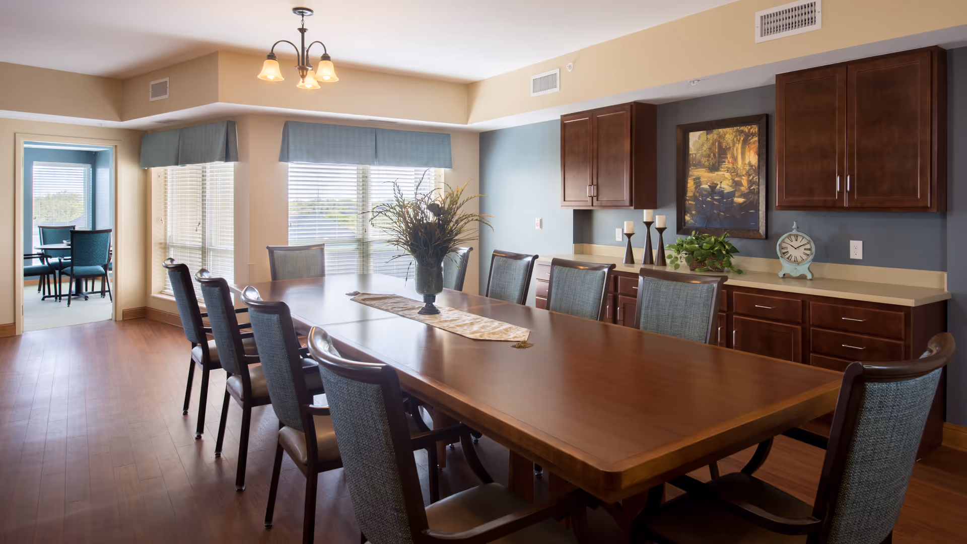 A spacious dining room with a long wooden table surrounded by ten chairs. The room has large windows with blinds and blue valances, allowing natural light to fill the space. On the right wall, there are wooden cabinets, a countertop with decorative items including candles, a clock, and a framed painting. In the background, there is an adjoining room with a smaller table and chairs near more windows.