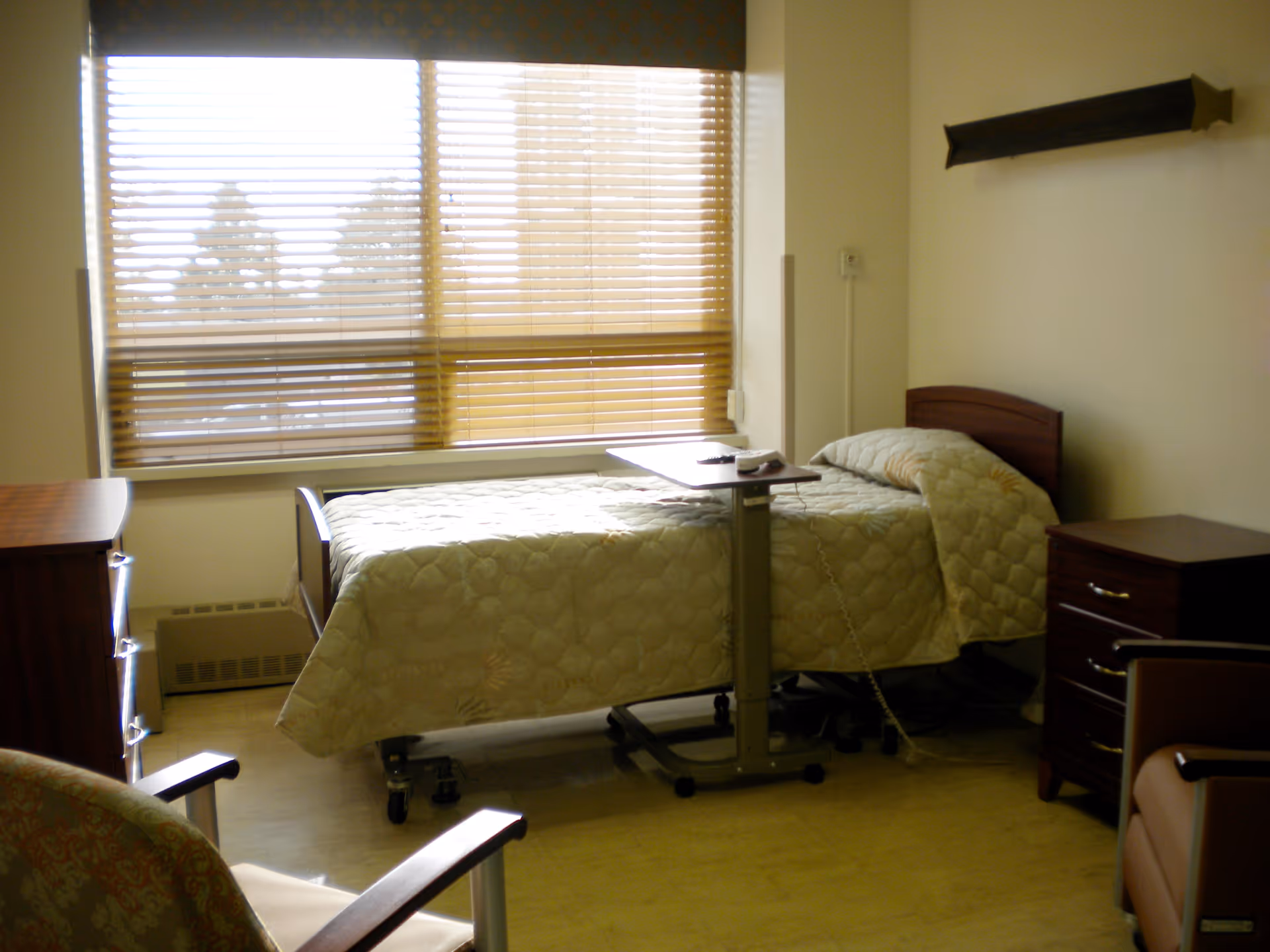 A simple senior-living bedroom with a single adjustable bed, overbed table, dresser, nightstand and chairs by a large window with blinds.