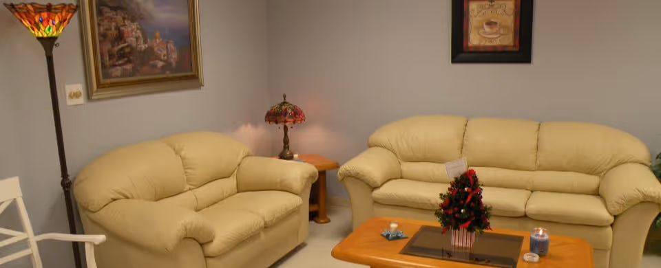 A cozy living room with two beige leather sofas, a wooden coffee table with a small decorated Christmas tree and candles, a side table with a stained glass lamp, a tall floor lamp with a colorful stained glass shade, and framed artwork on the light gray walls.