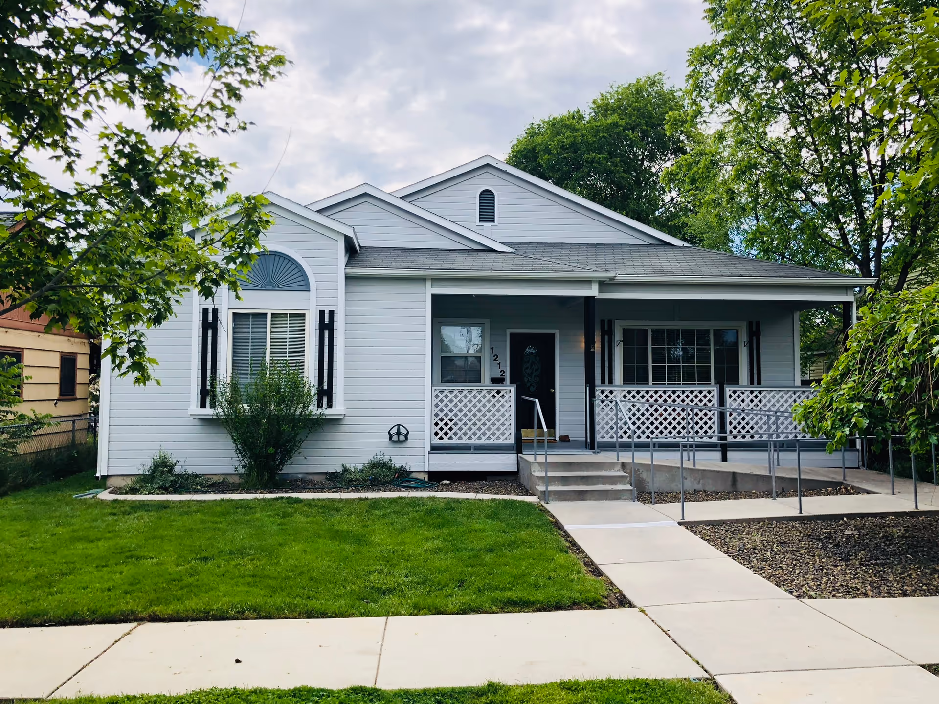 Front view of a single-story light-gray house with a covered porch, wheelchair ramp, steps, and a green lawn.