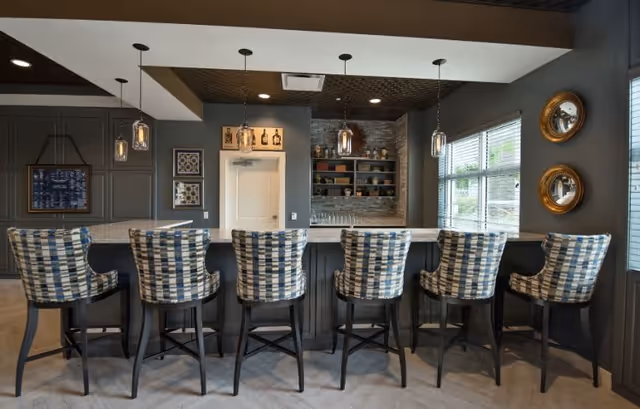 Interior view of a modern bar area with six high-back stools featuring a blue and white checkered pattern. The bar has a white countertop and dark paneling. Above the bar hang five pendant lights with glass shades. Behind the bar is a wall with shelves holding various decorative items and glassware. Large windows on the right side allow natural light to enter the room, and two round mirrors are mounted on the wall beside the windows.