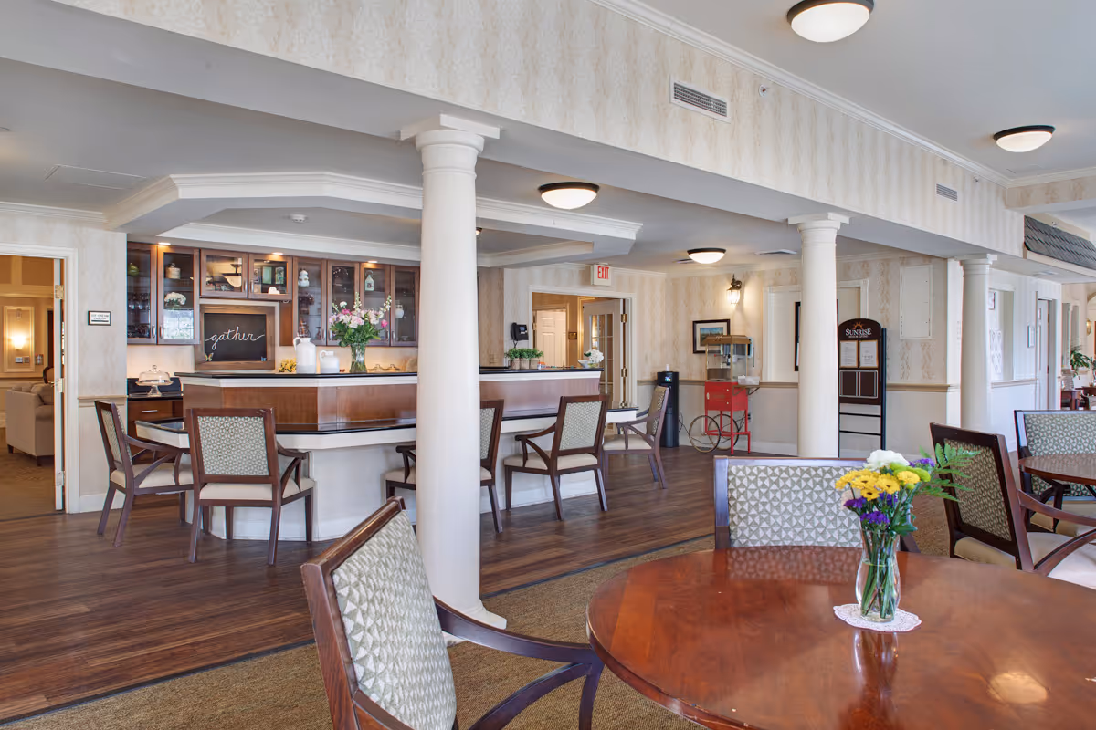 A spacious and well-lit common area in a senior living facility featuring a wooden round table with a vase of colorful flowers in the foreground, several chairs with patterned upholstery, white columns, a wooden reception or bar counter with glass cabinets behind it, and a popcorn machine near the back wall. The room has warm wooden flooring and soft beige wallpaper with subtle patterns.