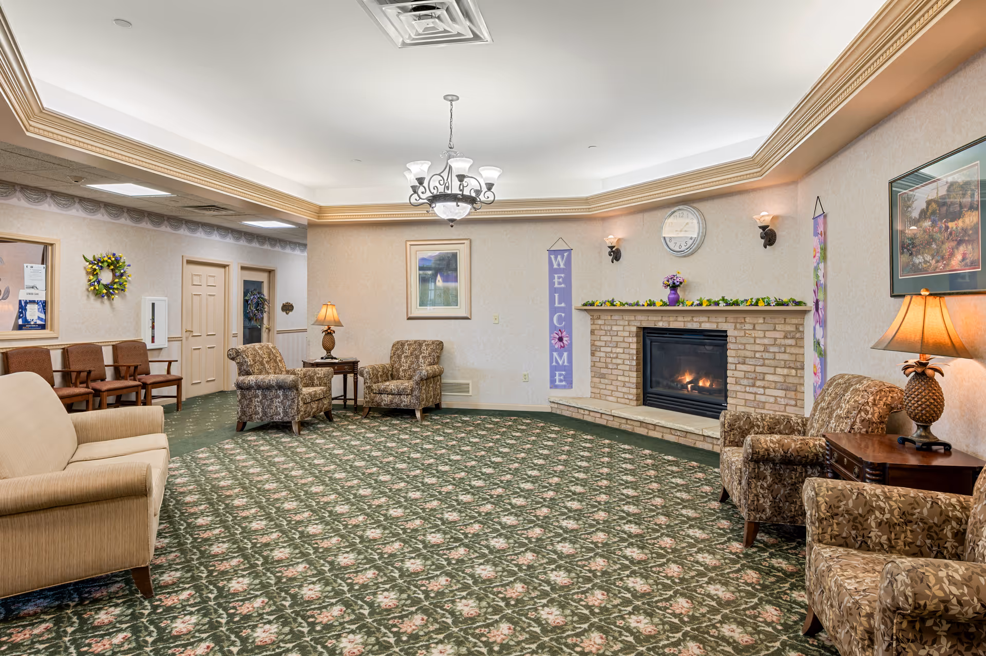 A cozy senior living facility common area with floral patterned green carpet, beige walls, and a brick fireplace with a fire burning. The room has several upholstered armchairs and a beige sofa arranged around the fireplace. There are two table lamps, a wall clock above the fireplace, a framed picture, and a vertical 'WELCOME' banner hanging on the wall. The ceiling features crown molding and a chandelier light fixture.