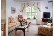 A cozy living room with a beige sofa, a floral armchair, a wooden coffee table with a flower arrangement, a leather recliner chair, a TV on a wooden stand, and a large window with decorative curtains letting in natural light.