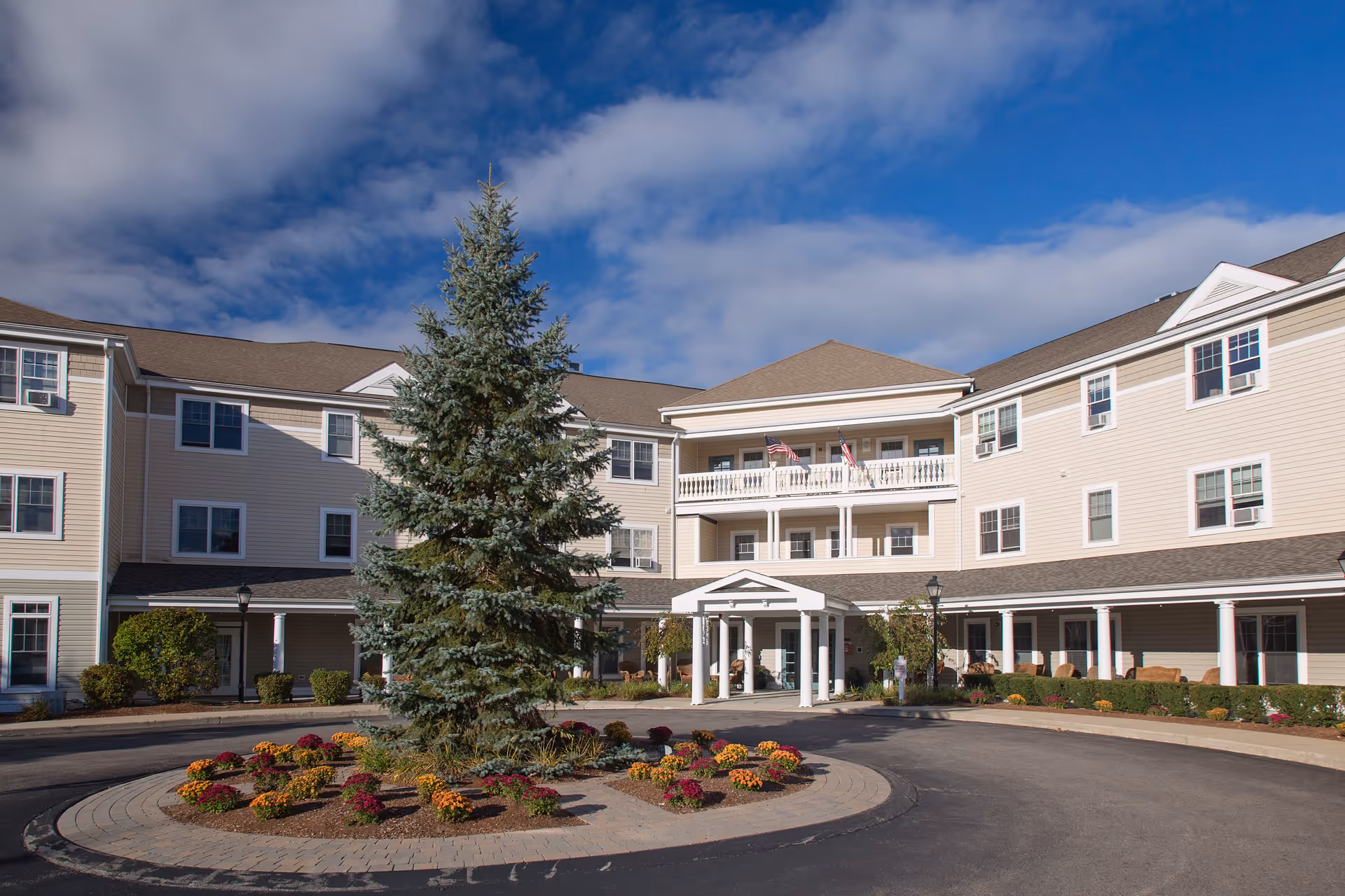 Exterior view of Traditions of Dedham senior living facility showing a three-story beige building with white trim, a circular driveway with a landscaped island featuring a large evergreen tree and colorful flowers, under a partly cloudy blue sky.