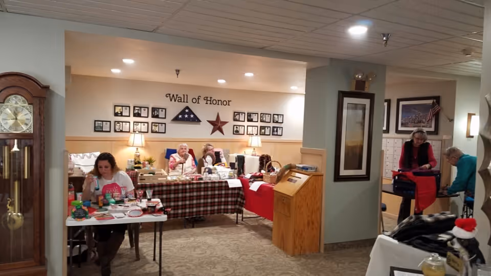 Interior view of a senior living facility room with several elderly people sitting and engaging in activities at tables covered with checkered and red tablecloths. The wall behind them features a 'Wall of Honor' with framed photos, a folded American flag in a triangular display case, and a large star decoration. The room has a grandfather clock on the left and framed artwork on the walls. Two people are standing and working on something at a table on the right side of the image.