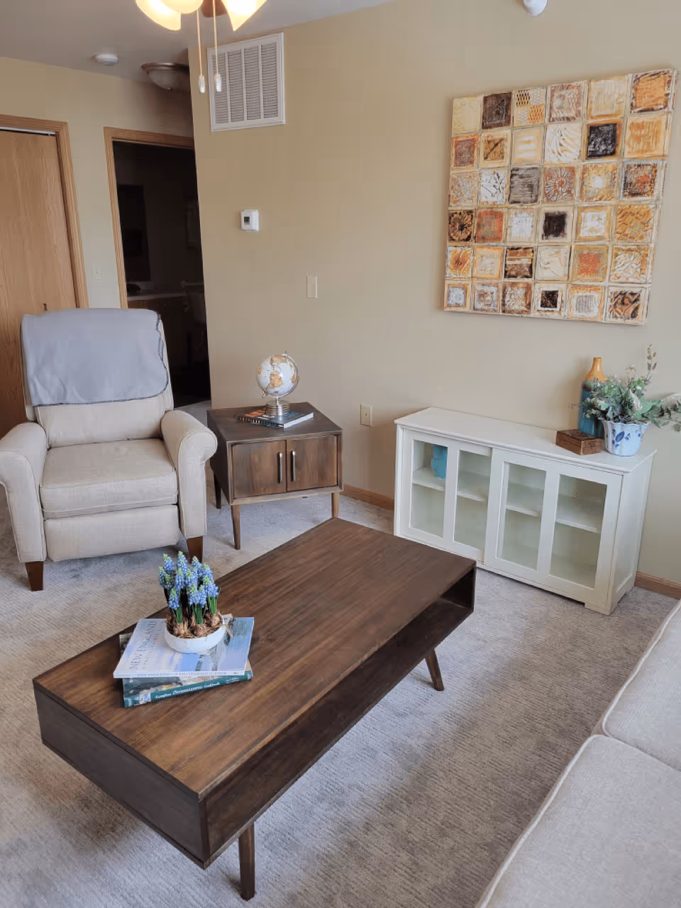 A cozy living room area with a beige armchair covered with a gray throw, a wooden side table with a globe and books, a wooden coffee table with a small potted plant and books, a white cabinet with glass doors, and a decorative wall art piece composed of various textured squares in warm tones.