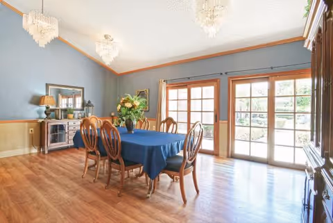 Dining room with a blue-covered wooden table and six chairs, chandeliers, and large glass French doors overlooking a garden.