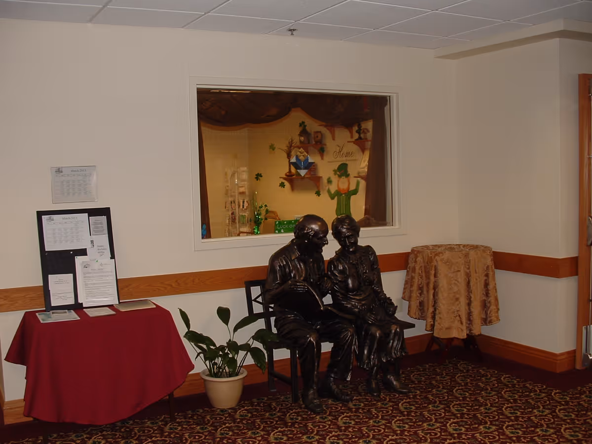 Interior corner of a room with a bronze statue of an elderly couple sitting on a bench. To the left is a table covered with a red cloth holding papers and a bulletin board with notices. A potted plant is on the floor next to the table. On the right side, there is a small round table covered with a gold patterned cloth. A window in the wall shows a decorated area with a leprechaun figure and the word 'Home'.