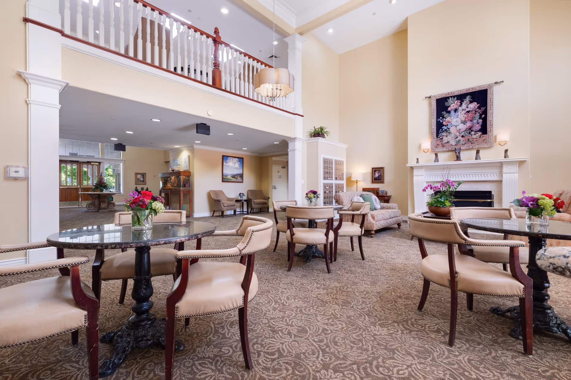 A spacious and elegant common area in a senior living facility featuring multiple round tables with beige cushioned chairs, floral centerpieces, a patterned carpet, a white fireplace with a floral tapestry above it, and a second-floor balcony with white railings. The room is well-lit with natural light coming through the entrance doors and ceiling lights.