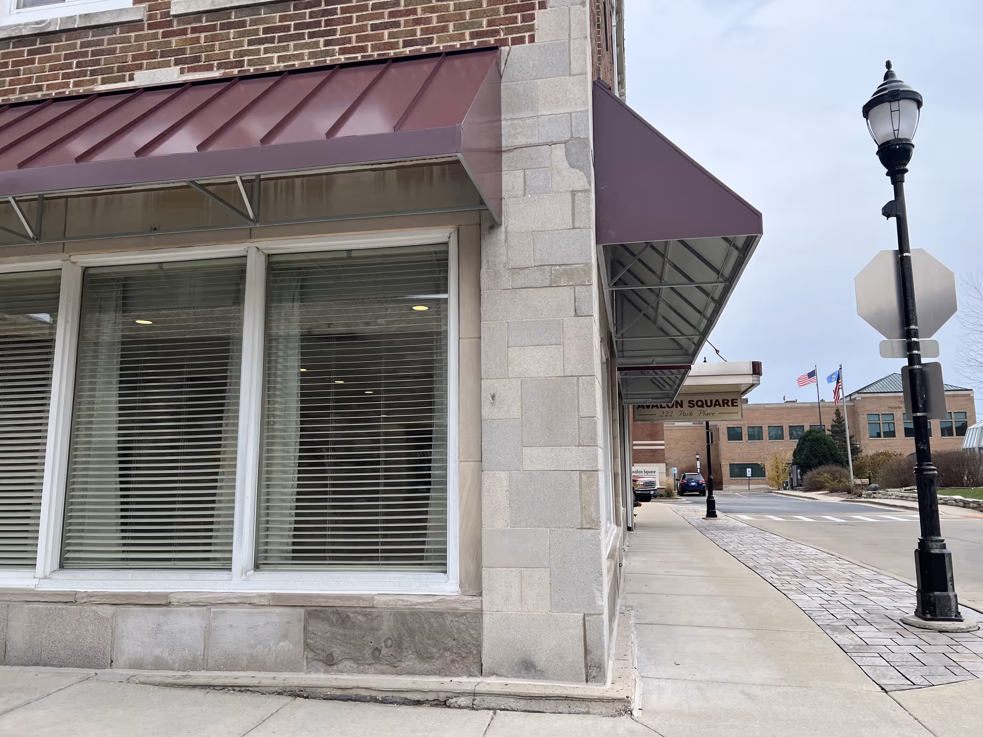 Street view of the exterior corner of a building with large windows covered by blinds and a maroon awning. A sidewalk runs alongside the building, leading to an entrance with a sign that reads 'Avalon Square'. There are street lamps and flags visible in the background under a cloudy sky.