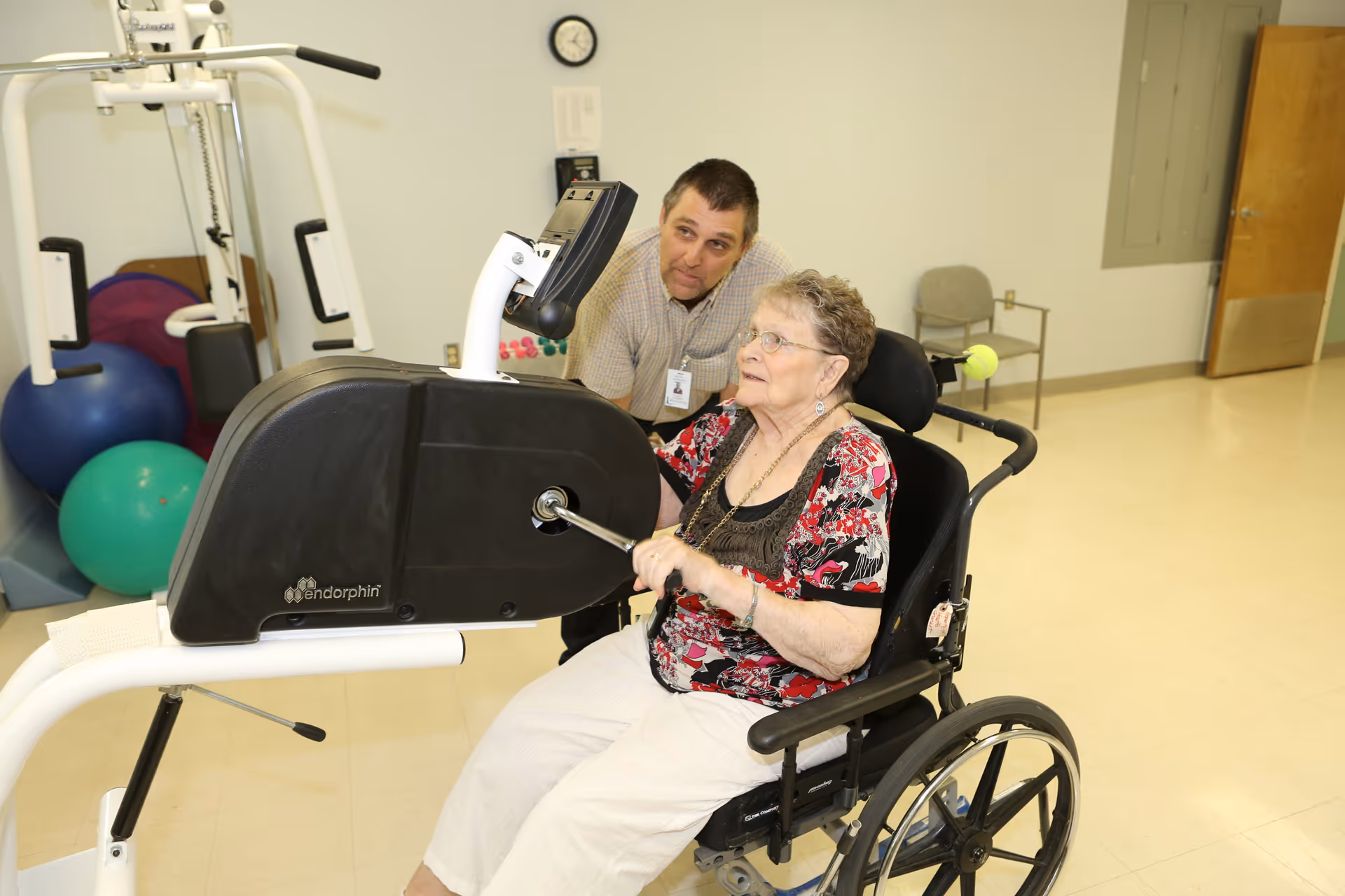 An elderly woman in a wheelchair is using a hand crank exercise machine in a rehabilitation or therapy room, assisted by a male staff member who is watching and supporting her. The room has exercise balls and other equipment in the background.