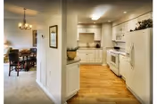 View of a kitchen with white cabinets, white appliances including a refrigerator, stove, and microwave. The kitchen has wooden flooring and is adjacent to a dining area with a table and chairs visible through an open doorway.