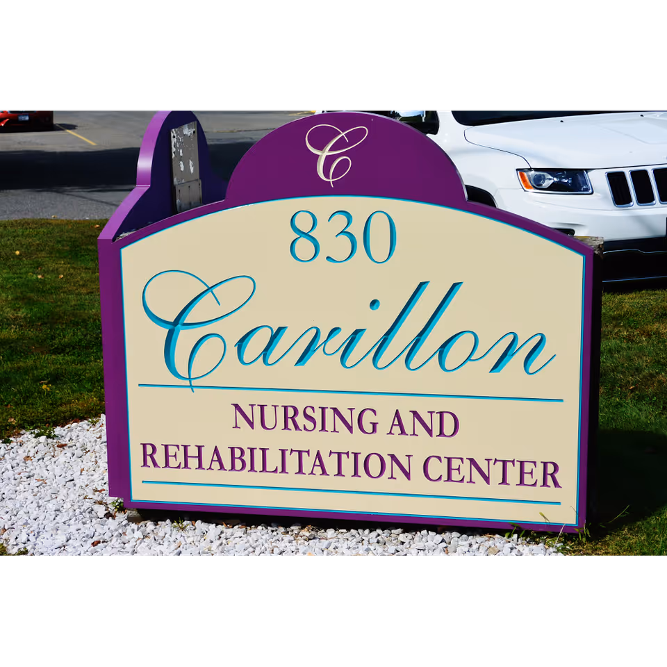 A purple and cream-colored sign for Carillon Nursing and Rehabilitation Center with the address number 830, placed on a grassy area with white gravel around its base. A white vehicle is partially visible in the background.