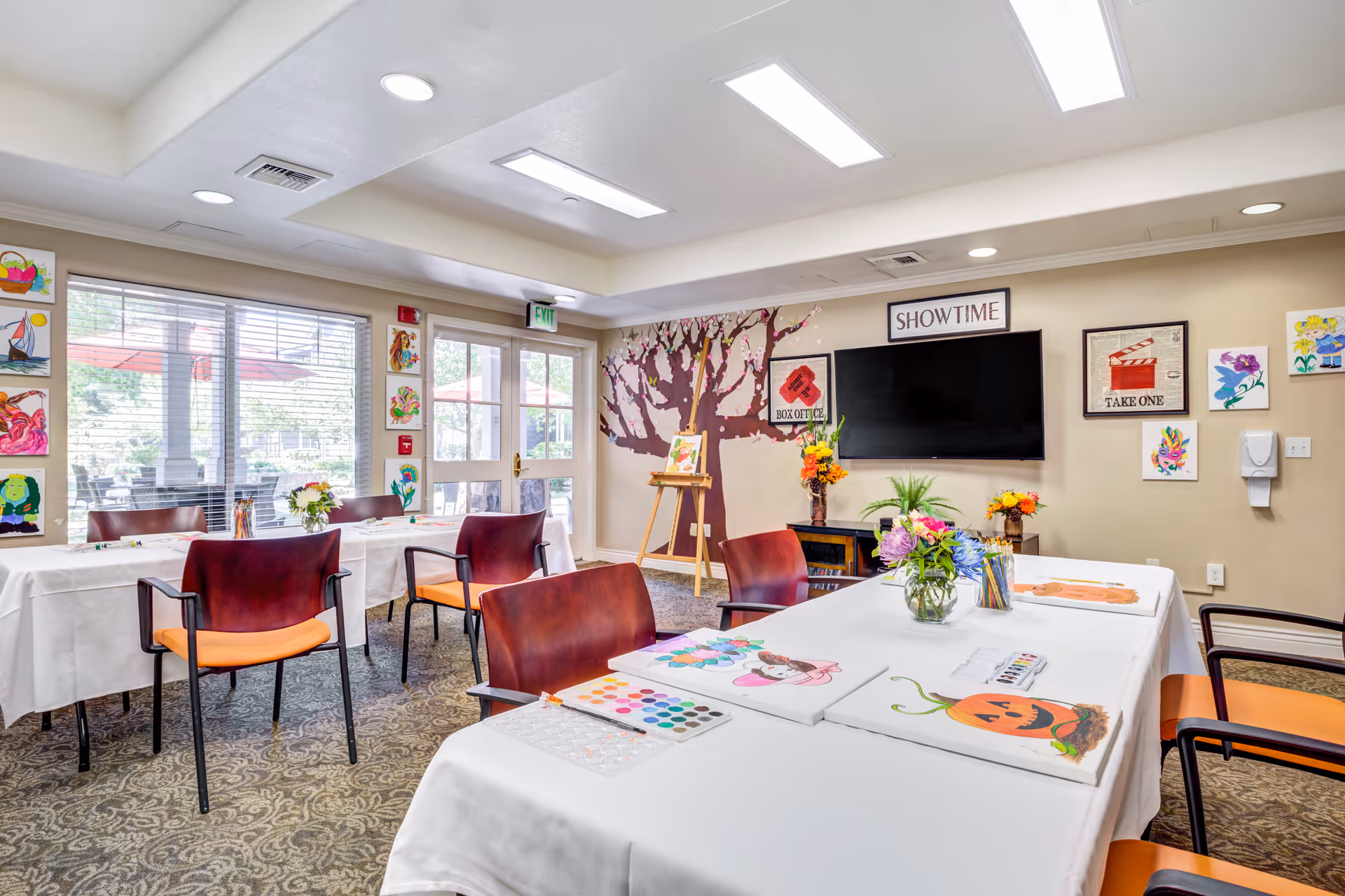 A bright and colorful activity room with tables covered in white cloths, set up for painting with watercolor palettes, brushes, and paintings of various subjects including a pumpkin and a girl. The room has several chairs with orange cushions, a large TV mounted on the wall with a 'SHOWTIME' sign above it, and walls decorated with artwork and a painted tree mural. Large windows and glass doors let in natural light and show an outdoor patio area with red umbrellas.