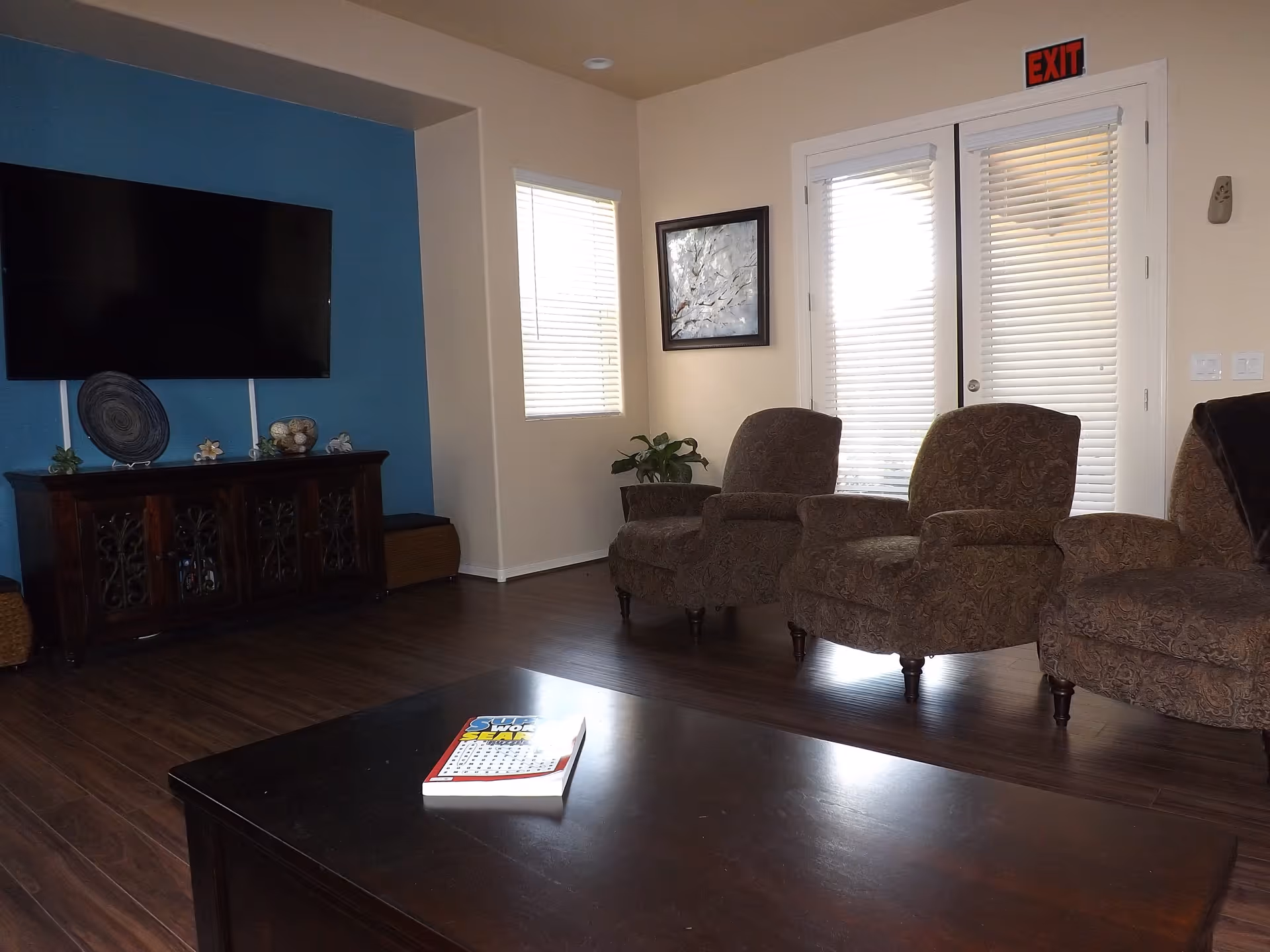 A living room with three patterned armchairs arranged near a set of French doors with blinds. A dark wooden coffee table with a book on it is in the foreground. A flat-screen TV is mounted on a blue accent wall above a dark wooden cabinet with decorative items. The floor is dark wood, and there is an exit sign above the doors.