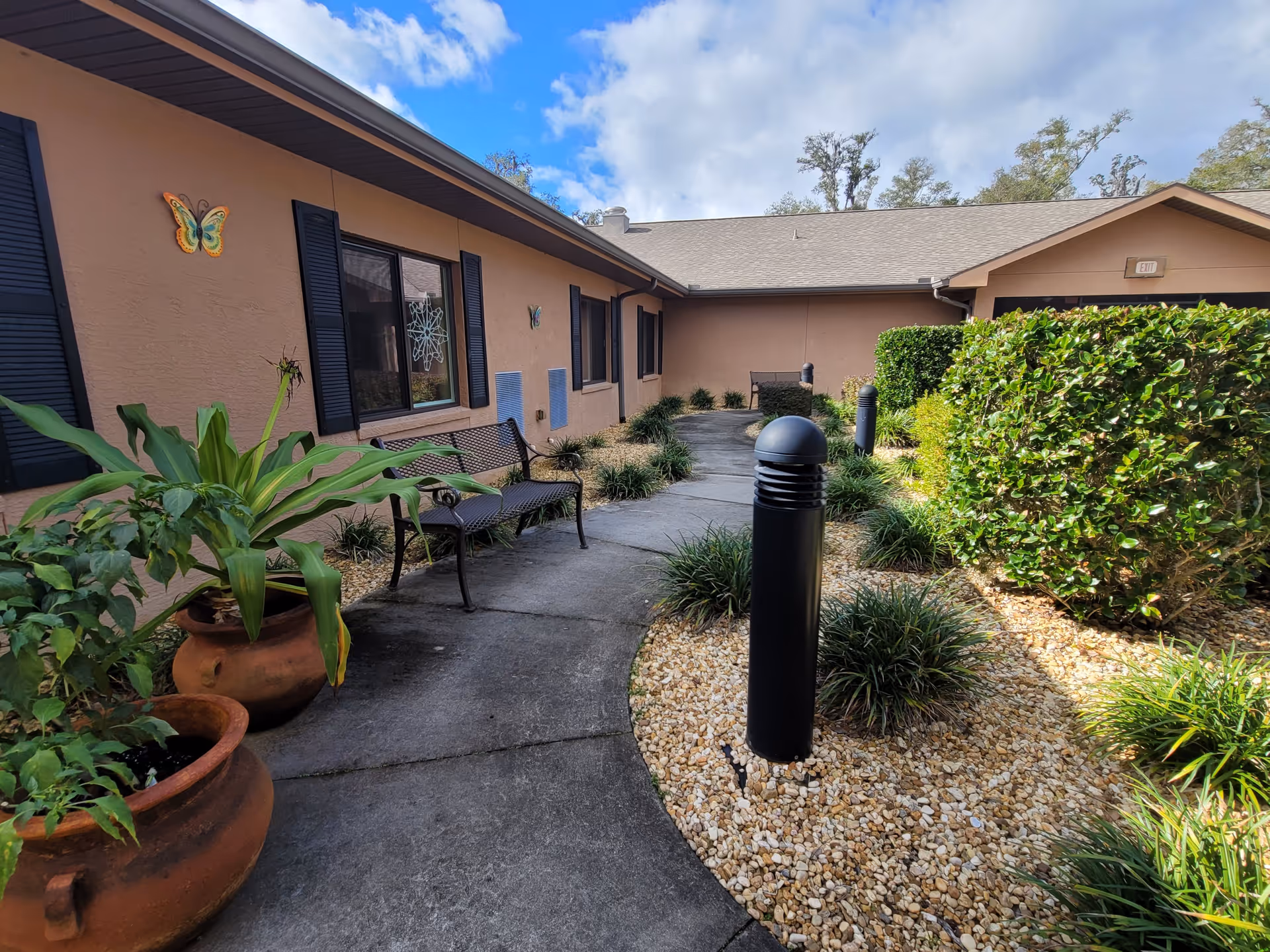 Outdoor courtyard area of a senior living facility with a curved concrete walkway, potted plants, bushes, and a bench along the building wall. The building has tan walls with black shutters and butterfly decorations. The sky is partly cloudy.