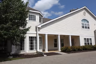 Front exterior of a two-story white assisted living building with a covered entrance supported by columns, multiple windows, and a driveway under a partly cloudy sky.