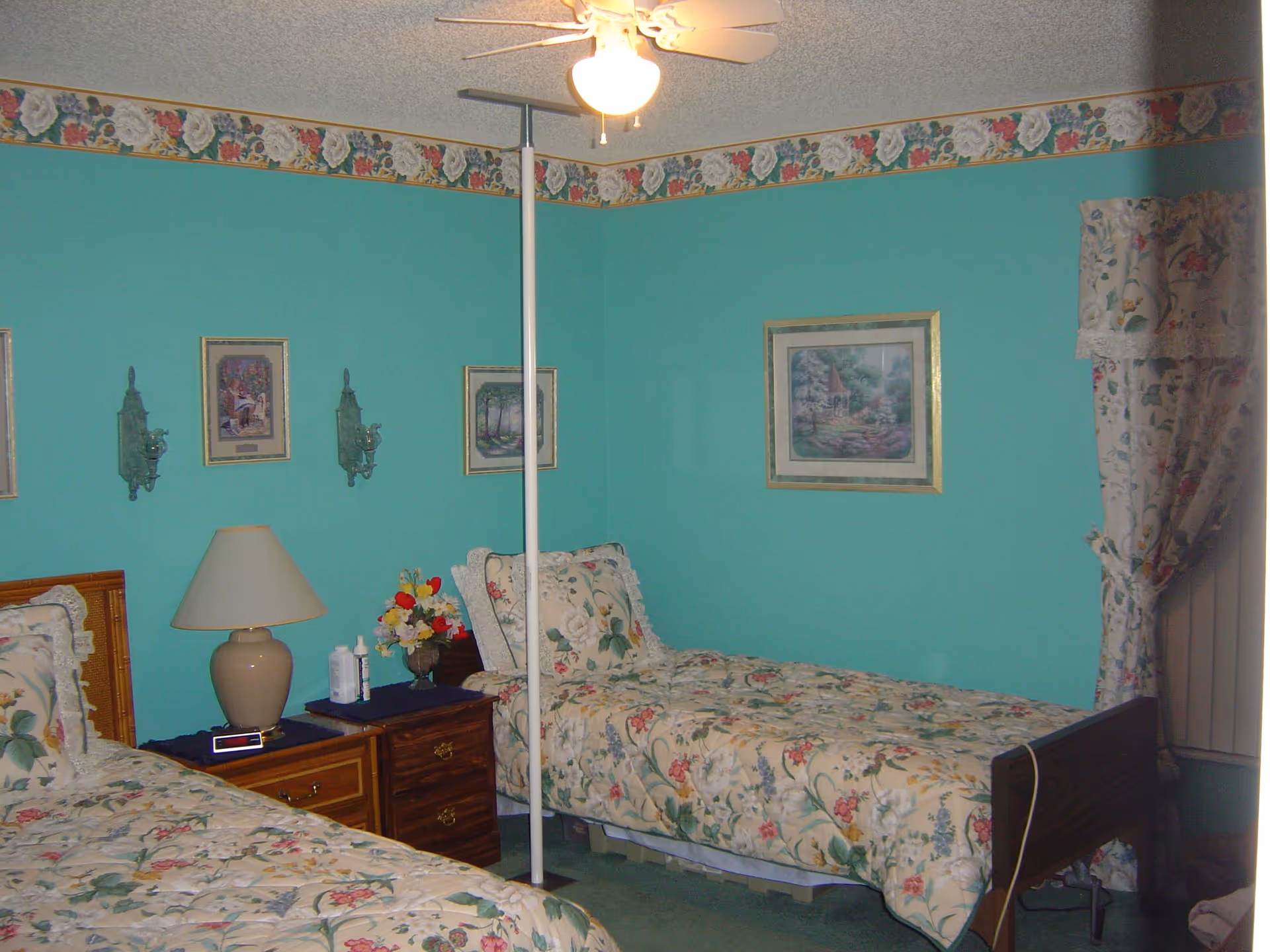A bedroom with two single beds covered in floral patterned bedspreads. Between the beds is a wooden nightstand with a beige lamp, an alarm clock, and some bottles. The walls are painted teal with a floral wallpaper border near the ceiling. There are framed pictures and decorative wall sconces on the walls. A ceiling fan with a light is on the ceiling, and floral curtains cover a window on the right side.