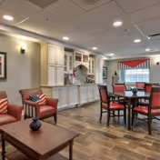 Interior view of a senior living facility common area with a seating arrangement including red cushioned chairs and a wooden coffee table. There is a dining table with chairs in the background near a window with red and white striped valance. The room features built-in white cabinetry and wood-look tile flooring under recessed ceiling lights.
