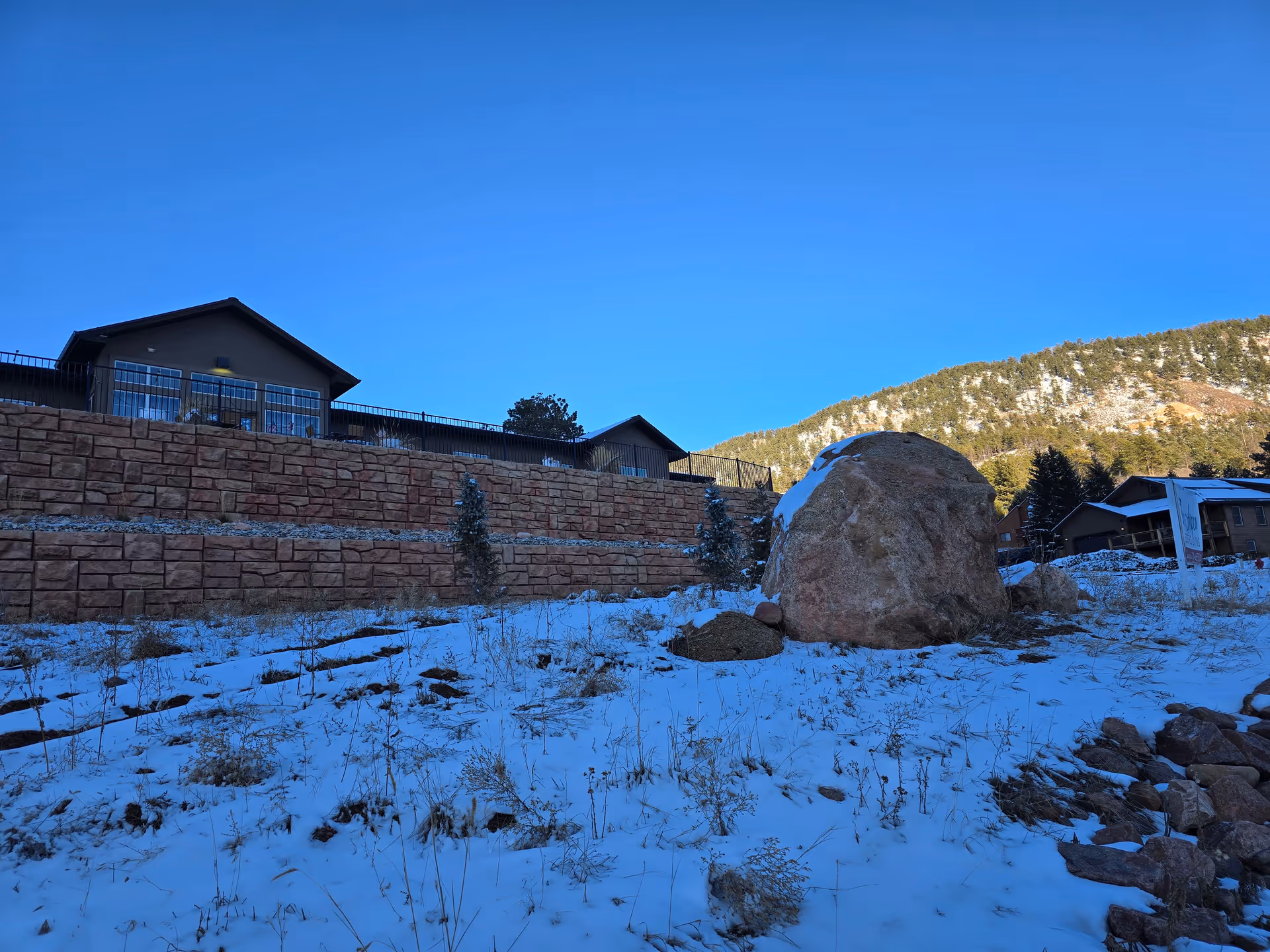 Snow-covered ground with dry grass and rocks in the foreground, a large boulder to the right, and a retaining wall with a building and railing above it. In the background, there is a forested hillside under a clear blue sky.